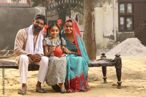 Happy rural Indian family sitting together, daughter is holding a piggy bank in her hand, putting a coin in it, showing the concept of saving money