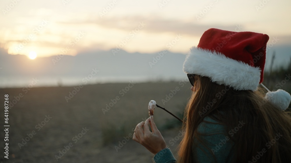 Obraz premium A young woman in a Santa hat on the background of mountains and sunset eats fried marshmallows on the beach by the sea. New Year.