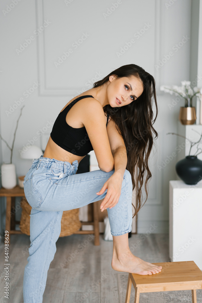 Sensual beautiful young Caucasian woman with long hair in a black top and jeans posing for the camera in the living room