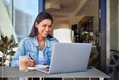 Young female student studying in cafe with laptop taking notes