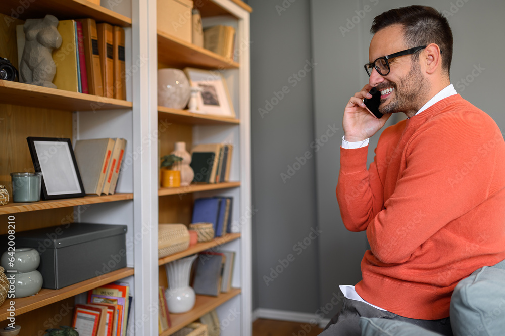 Side view of young businessman talking over smart phone while sitting in front of bookshelf at home Side view of young businessman talking over smart phone while sitting in front of bookshelf at home