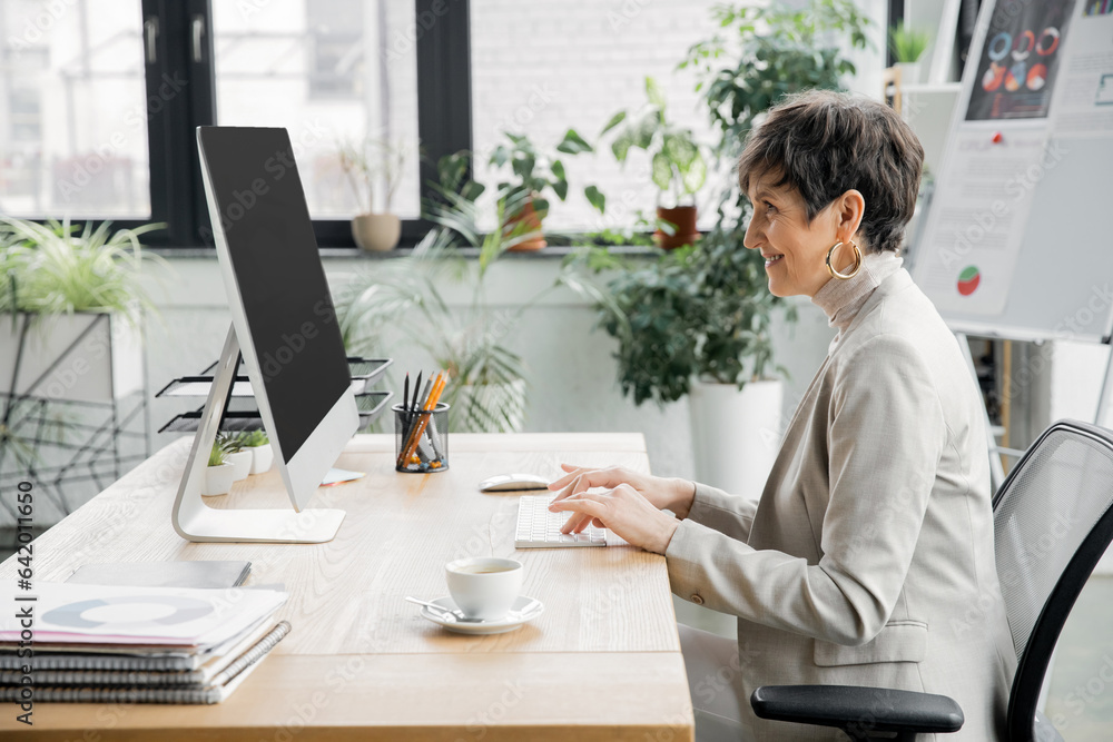 side view of middle aged businesswoman working on compute near coffee cup and documents in office side view of middle aged businesswoman working on compute near coffee cup and documents in office