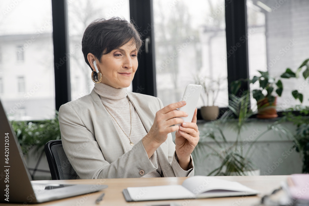 pleased mature businesswoman in earbud browsing internet on smartphone near laptop in office pleased mature businesswoman in earbud browsing internet on smartphone near laptop in office