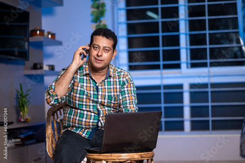 Technology, remote job and lifestyle concept - happy Indian man Talking on phone while working on laptop computer at home office.
