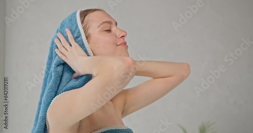 Attractive woman wrapped in towel drying blonde hair with towel after shower. Side view of relaxed caucasian girl wiping wet hair in bathroom, in motion. Concept of hair care routine.