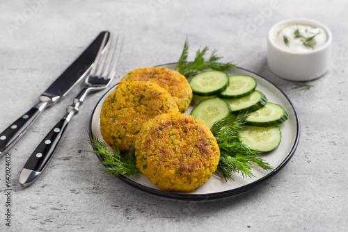 Delicious millet cutlets with carrots and seeds served with cucumber and herbs on a gray textured background, top view. Homemade vegan food