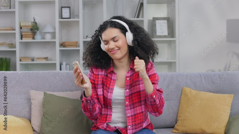 Charming african american young lady dancing with hands while sitting on grey sofa in headphones. Pretty woman with closed eyes enjoying favorite music from cellphone during free time at apartment. Charming african american young lady dancing with hands while sitting on grey sofa in headphones. Pretty woman with closed eyes enjoying favorite music from cellphone during free time at apartment.