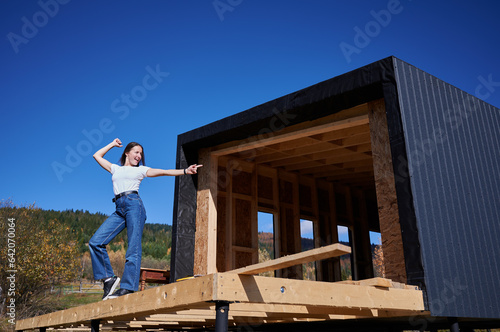 Happy woman on construction site standing on terrace at unfinished wooden frame house on sunny day, pointing at future building.