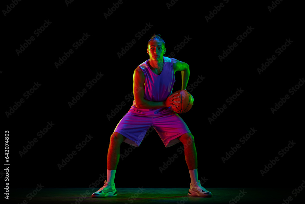 Young, active, motivated guy, basketball player in uniform standing in position with ball against black background in neon light. Concept of professional sport, competition, hobby, game, competition