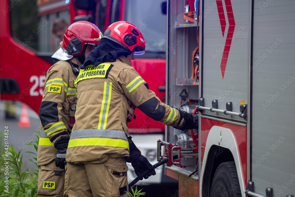 Obraz premium Strażacy podłączający wąż. Straż pożarna przy wypadku. Strażacy gaszą samochód. Straż pożarna gasi płonący samochód. Firemen connecting a hose. The fire department at the accident. Firefighters exting