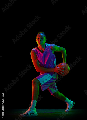 Full-length dynamic image of teen boy, basketball player in uniform with ball in motion against black studio background in neon light. Professional sport, competition, hobby, game, competition concept