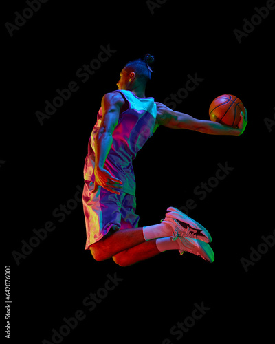 Athletic young guy, basketball player in motion during match, jumping with ball against black studio background in neon light. Concept of professional sport, competition, hobby, game, competition