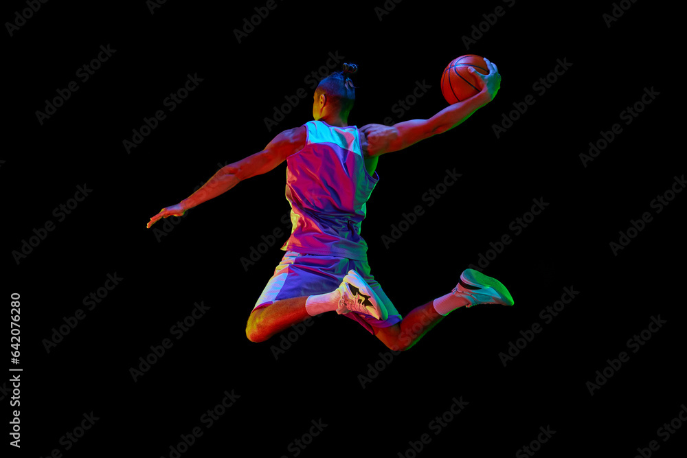 Back view image of young guy, basketball player in sportswear training, jumping and throwing ball against black studio background in neon light. Concept of sport, competition, hobby, game, competition