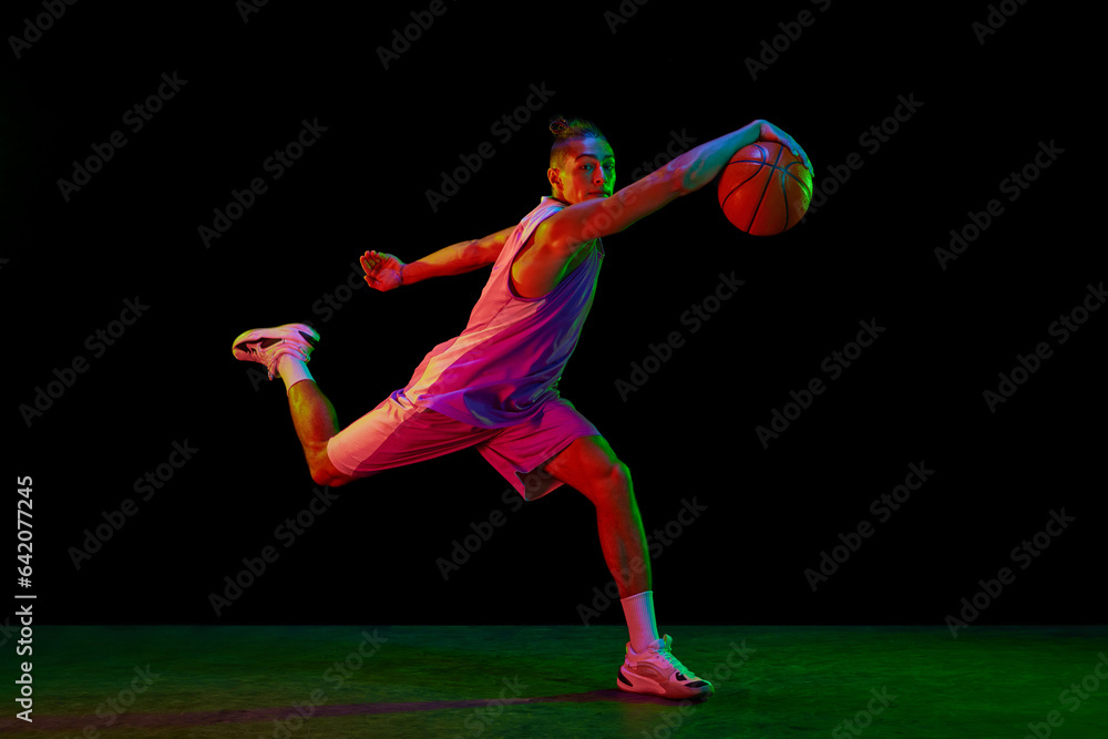 Muscular, active young man in motion, basketball player with ball during game against black background in neon light. Concept of professional sport, competition, hobby, active lifestyle, competition