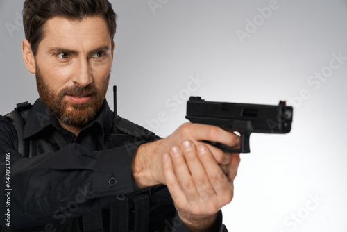 Focused bearded officer taking aim, by holding handgun both hands in studio. Portrait of caucasian cop keeping gun, zeroing in side, looking away, on gray background. Concept of danger work, weapon.