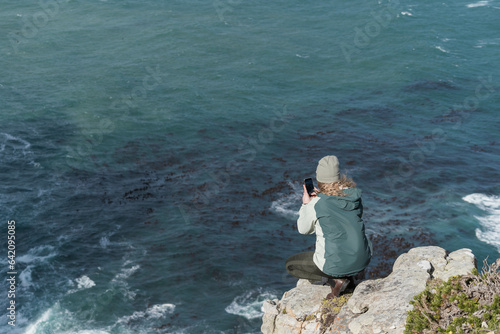 Woman taking a photo of the ocean from a phone whilst standing at the top of a cliff on a rock.