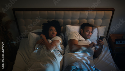 African American couple family in bed bedroom at night angry frustrated woman wife trying sleep offended on husband man gadget addict chatting with phone laughing mobile smartphone addiction problem