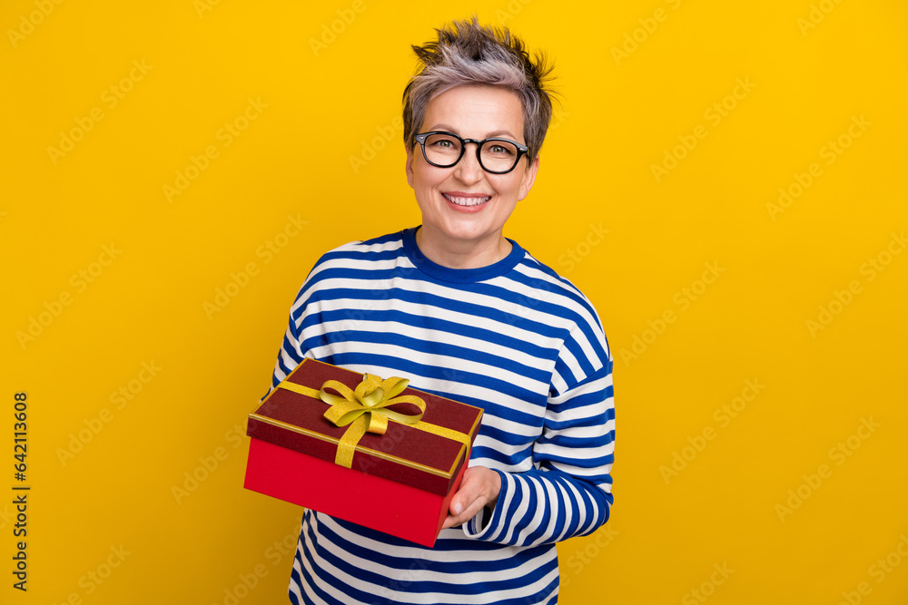 Photo of optimistic elderly grandmother wear striped trendy shirt hold red gift box happy birthday ad isolated on yellow color background Photo of optimistic elderly grandmother wear striped trendy shirt hold red gift box happy birthday ad isolated on yellow color background
