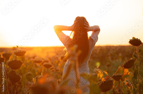 young woman in evening sunlight in a sunflower field at summer sunset, girl holding hands on hair standing back to the camera, relaxation on nature