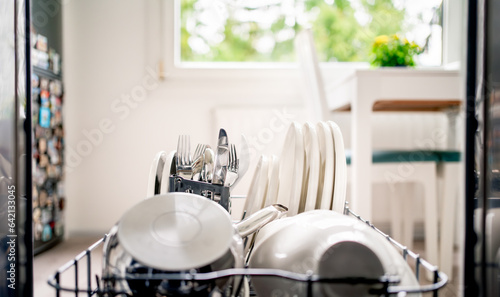 Dish washing machine. Inside an open dishwasher, view to kitchen. Clean plates, utensils, cutlery and pot in washer rack and shelf. Basket with fork and knife. Full load. Domestic appliance technology