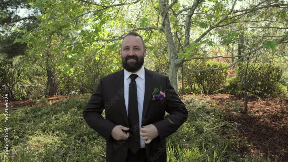 Middle-aged dark-haired unshaven groom putting on black jacket with boutonniere. Smiling groom in wedding suit standing outdoors among trees on sunny day. High quality 4k footage Middle-aged dark-haired unshaven groom putting on black jacket with boutonniere. Smiling groom in wedding suit standing outdoors among trees on sunny day. High quality 4k footage