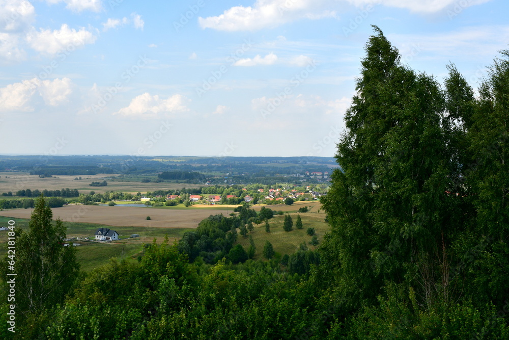 A close up on a hill covered with grass, trees and shrubs located next to a small village and close to some wind turbines and a tourist path seen on a sunny summer day in Poland during a hike