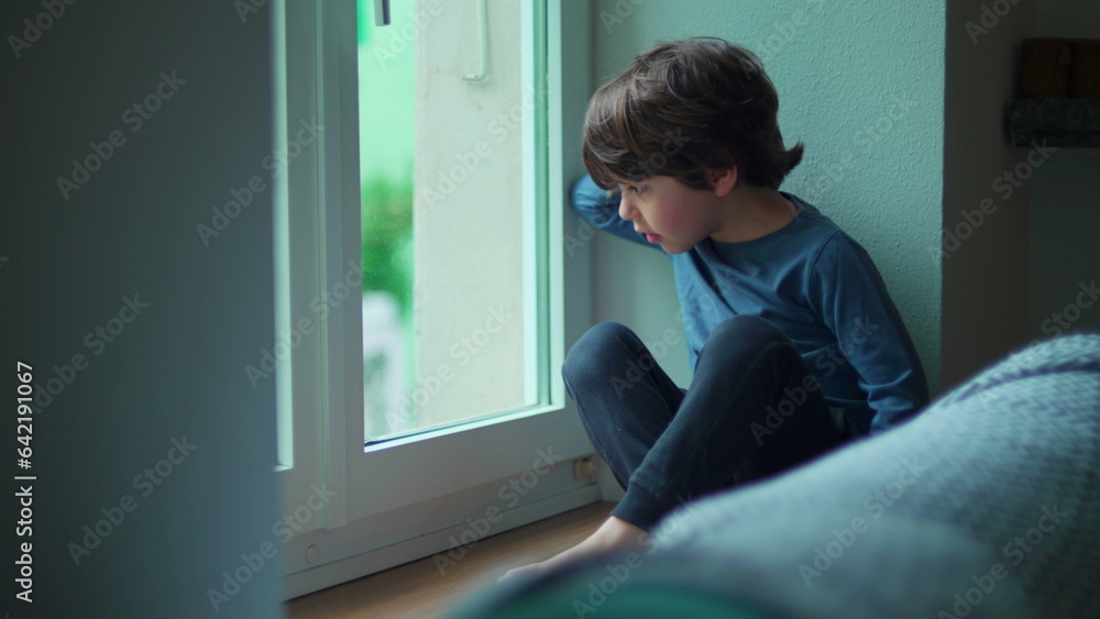 Child sitting by window at apartment staring out into street, leaning hand on glass looking at view from home Child sitting by window at apartment staring out into street, leaning hand on glass looking at view from home