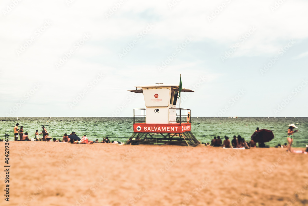 Fototapeta premium Bapselona, Spain. August 1, 2023 White lifeguard tower on sandy beach against green water in a sea. People relaxing on the Mediterranean coast on summer vacation in summertime Tilt shift travel photo.