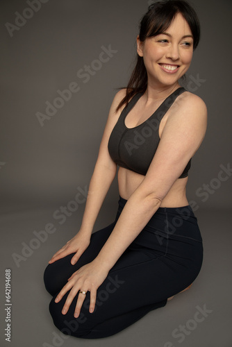 Studio portrait of a caucasian woman wearing a sports bra and yoga pants. She is smiling and sitting on the floor. She has her dark hair in a pony tail. 
