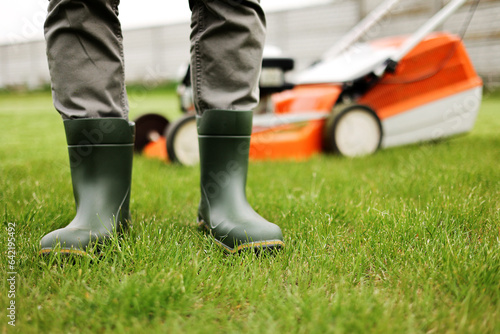Cropped photo of male gardener legs in rubber boots stands on cut green grass lawn at backyard of house. Modern electric cordless lawnmower on back. Landscaping industry theme.