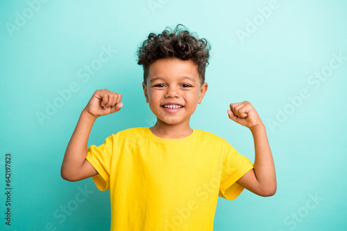 Portrait of good mood strong small boy with curly hair wear stylish t-shirt clenching fists isolated on turquoise color background