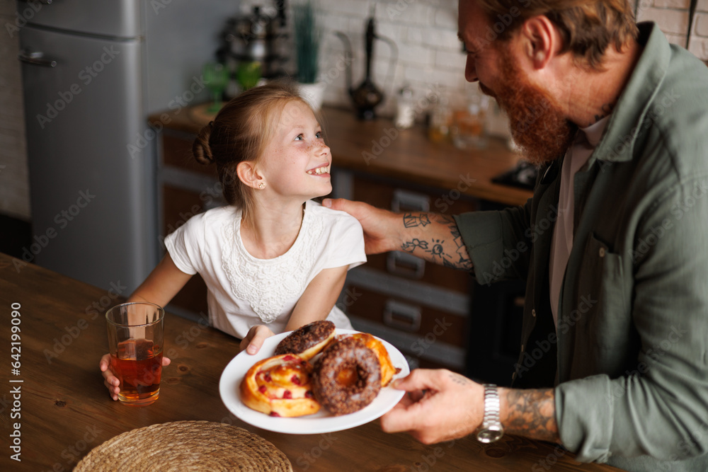 Tattooed father holding sweet pastry and talking to smiling daughter with juice in kitchen in morning