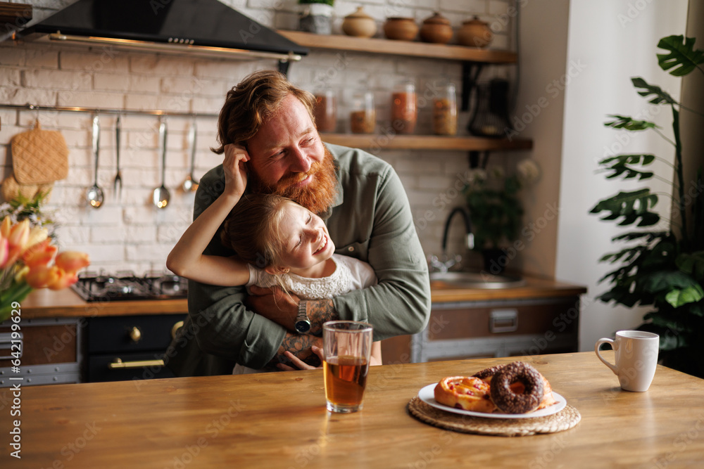 Cheerful daughter hugging bearded father near sweet breakfast in kitchen in morning - obrazy, fototapety, plakaty 