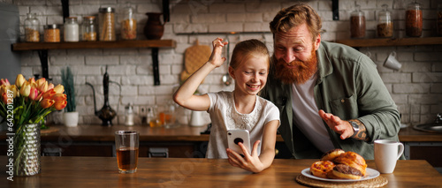 Banner image of cheerful bearded father and preadolescent daughter using smartphone together near pastry in kitchen 