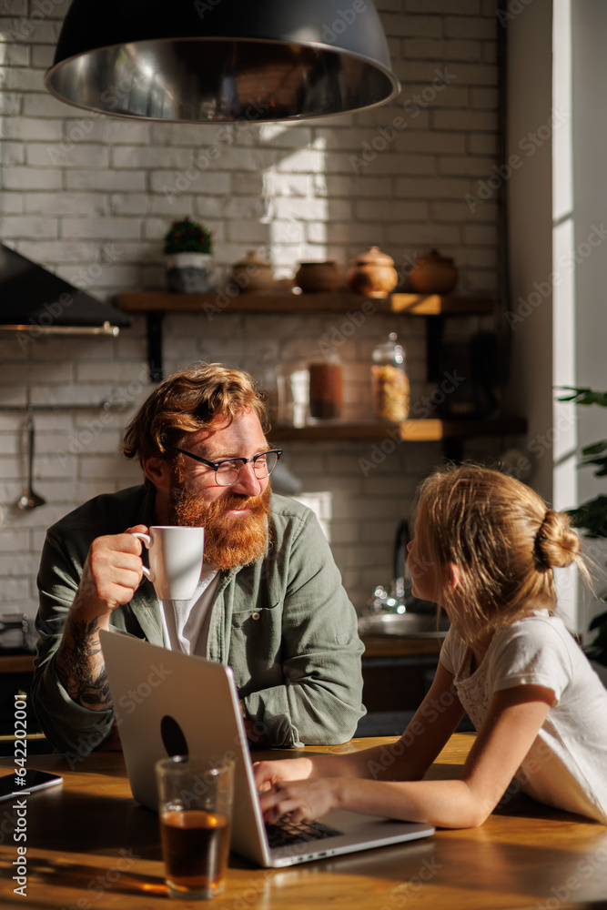 Smiling father holding coffee cup while daughter using laptop in kitchen in morning Smiling father holding coffee cup while daughter using laptop in kitchen in morning