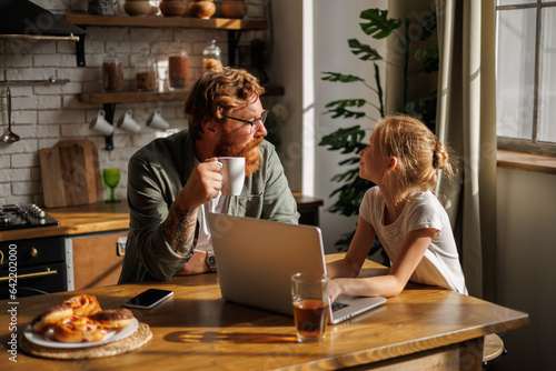 Father holding coffee cup and talking to smiling daughter using laptop during breakfast in kitchen 
