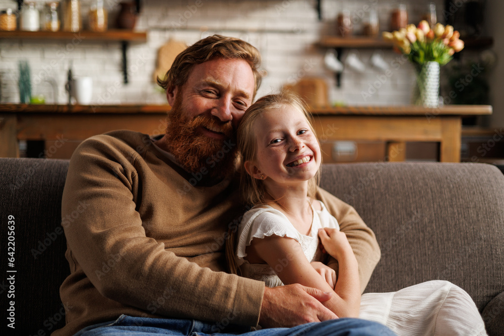 Portrait of smiling father hugging daughter and looking at camera on couch at home Portrait of smiling father hugging daughter and looking at camera on couch at home