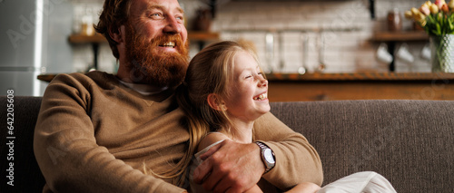 Banner image of smiling bearded father hugging preadolescent daughter on couch at home
