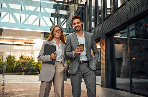 Creative modern stylish confident elegant beautiful successful cheerful carefree business woman and man standing outside business building holding laptop and coffee and looking at camera smiling. photo