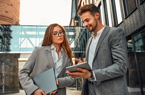 Creative modern stylish confident elegant business woman and man working together developing a project. A man shows a document on a tablet to a colleague. In the background, a business building. photo