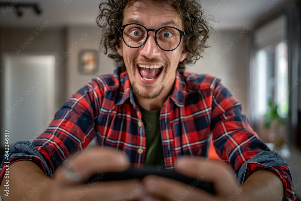 One adult caucasian man sit at home happy smile play video games leisure activity having fun hold mobile phone smartphone have fun copy space wear eyeglasses and shirt One adult caucasian man sit at home happy smile play video games leisure activity having fun hold mobile phone smartphone have fun copy space wear eyeglasses and shirt