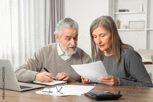 Elderly couple with papers and laptop discussing pension plan at wooden table in room