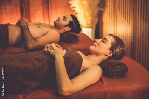 Caucasian couple customer enjoying relaxing anti-stress spa massage and pampering with beauty skin recreation leisure in warm candle lighting ambient salon spa at luxury resort or hotel. Quiescent