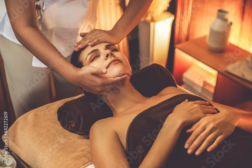 Caucasian woman enjoying relaxing anti-stress head massage and pampering facial beauty skin recreation leisure in warm candle lighting ambient salon spa in luxury resort or hotel. Quiescent