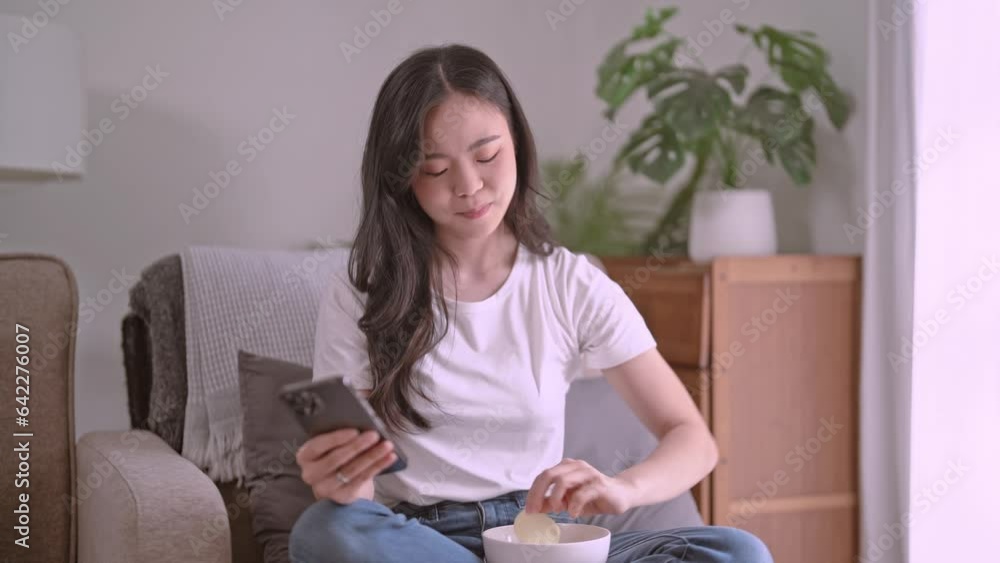Asian woman eating potato chips while using mobile phone to watching variety show and relax sits at home. Asian woman eating potato chips while using mobile phone to watching variety show and relax sits at home.