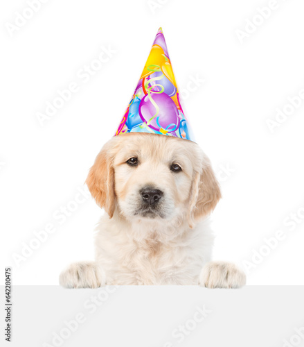 Golden retriever puppy wearing party cap looks above empty white banner. isolated on white background © Ermolaev Alexandr