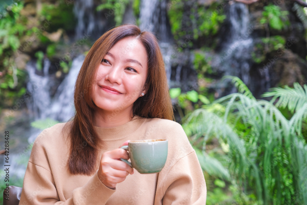 Portrait image of a young woman holding and drinking coffee while sitting in the garden with waterfall Portrait image of a young woman holding and drinking coffee while sitting in the garden with waterfall