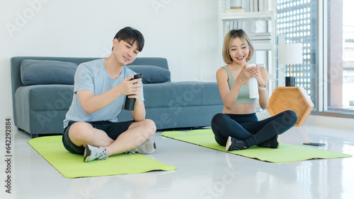 Asian young fit male female husband and wife in sportswear sport bra legging sneakers sitting smiling on yoga mat floor taking coffee break drinking water from bottles talking together in living room