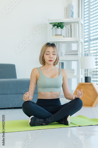 Asian young fit female teenager in sportswear sport bra legging sneakers sitting closed eyes meditating on yoga mat with lotus posture exercising training together in living room at home.