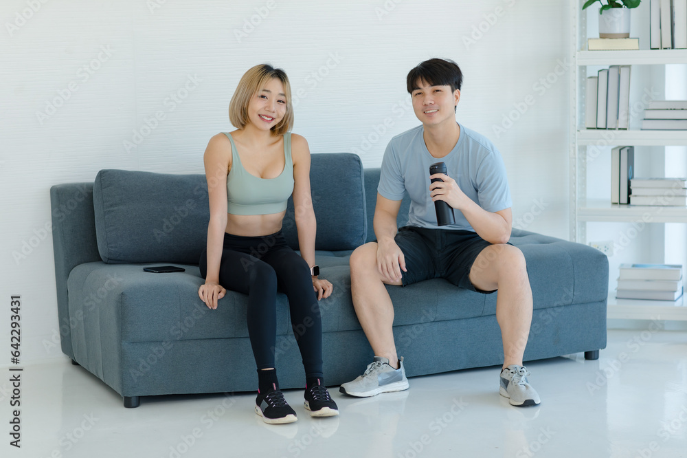 Obraz premium Asian young fit male female husband wife in sportswear sport bra leggings sneakers sitting on sofa talking smiling working out practicing exercising training together in living room at home